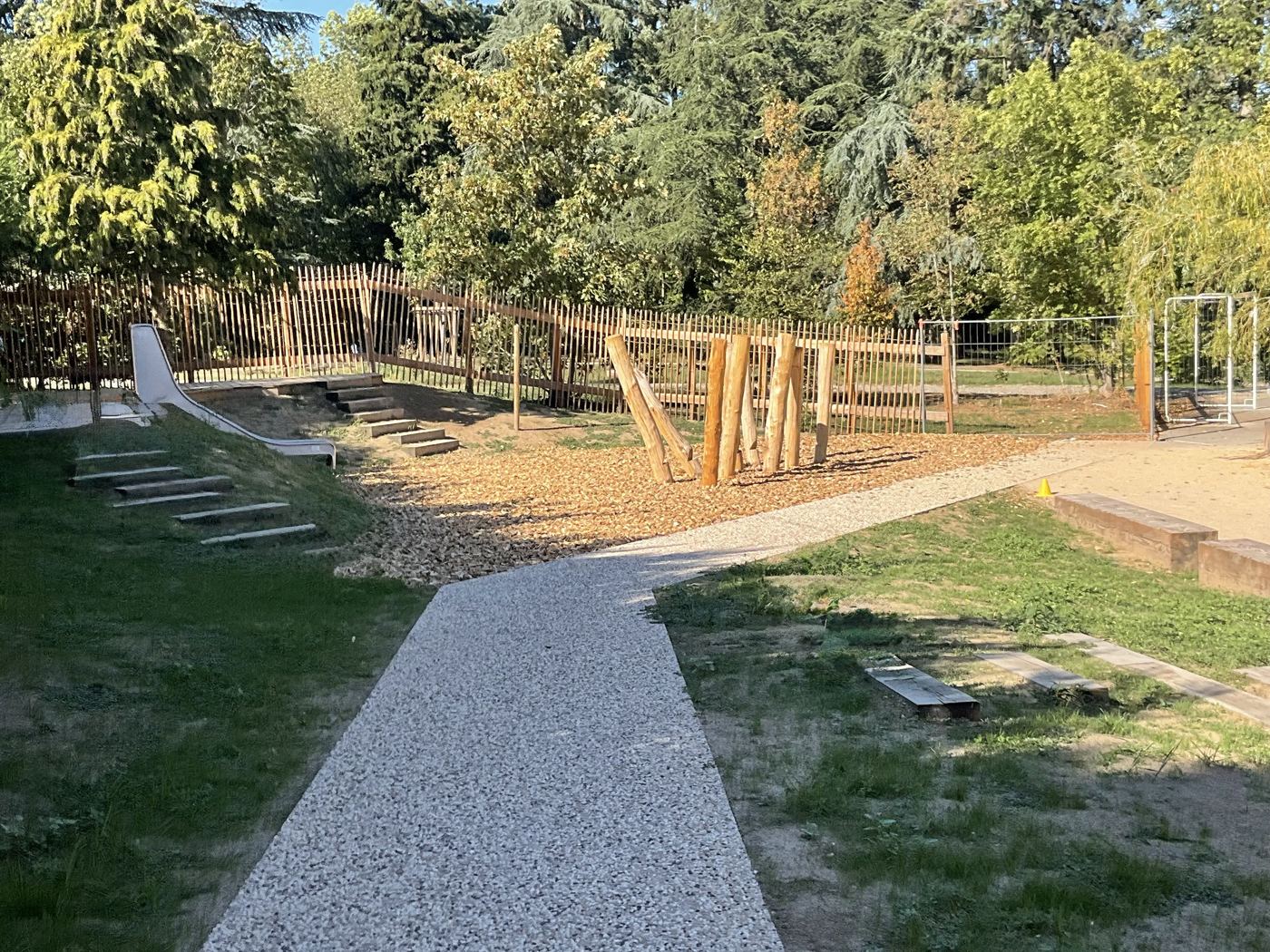 Le cheminement sous les arbres : bancs en pierre, ombres allongées des troncs sur l'allée.